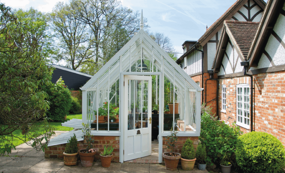 A white, Victorian-style greenhouse with attached cold frames on a red brick base.