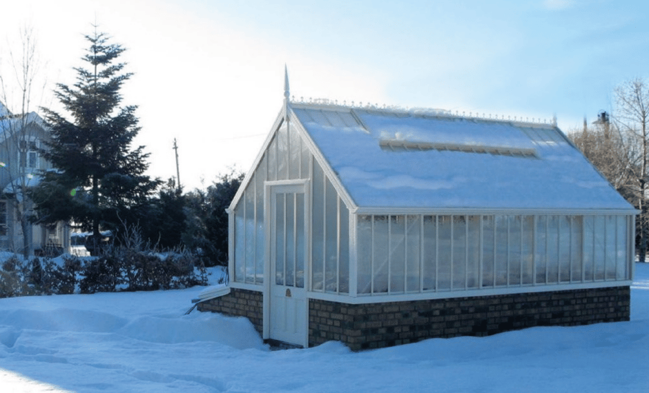 A victorian greenhouse in white, slightly buried in snow and on a brick base.