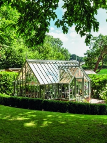 Alitex Tatton Victorian greenhouse with a lobby and vents. It is in a bright garden, with green foliage and trees.