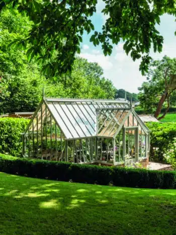 Alitex Tatton Victorian greenhouse with a lobby and vents. It is in a bright garden, with green foliage and trees.