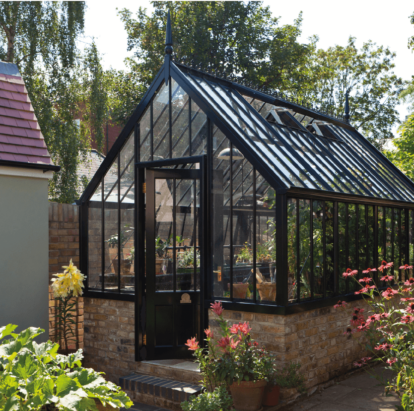 A victorian-style aluminium greenhouse in black, on a raised brick base. It is surrounded by green foliage and colourful plants.