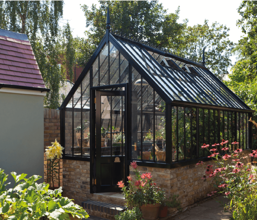 A victorian-style aluminium greenhouse in black, on a raised brick base. It is surrounded by green foliage and colourful plants.