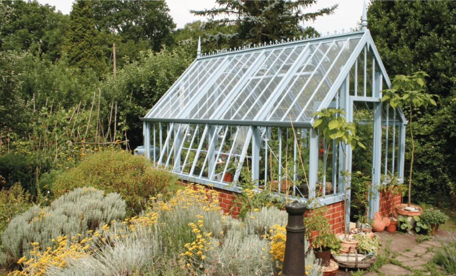 Light blue victorian-style Greenhouse with open side-vents on a red brick base. It is surrounded by green foliage and trees.