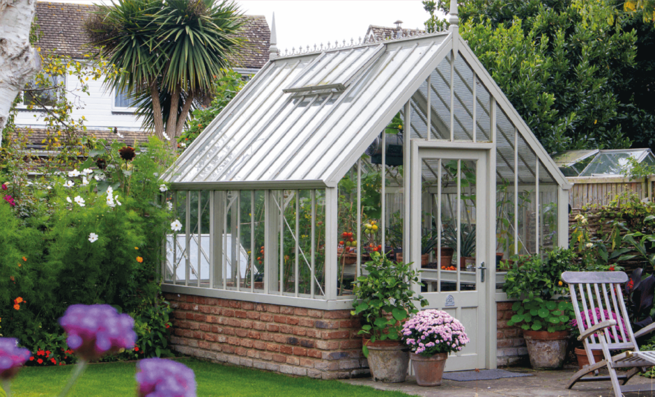 Victorian-Style Hidcote Greenhouse in White on top of a red brick base. The greenhouse is surrounded by colourful potted plants and nestled in the corner of a garden.
