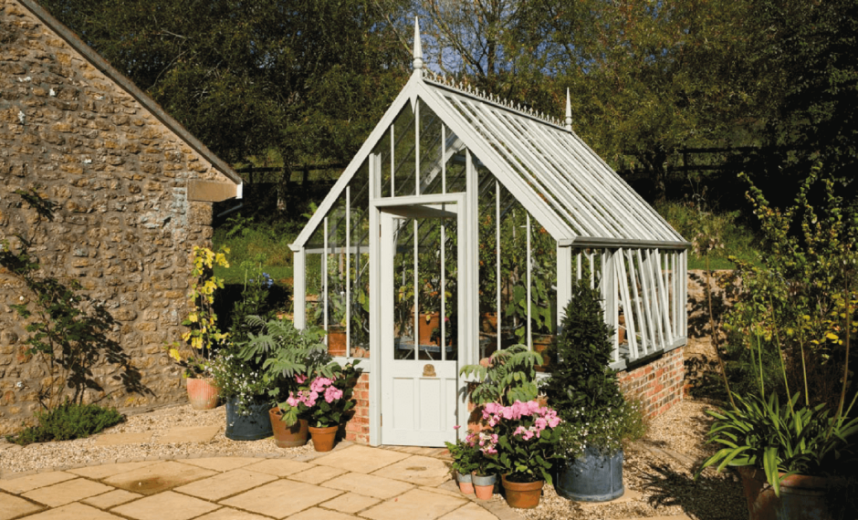 White Victorian-style greenhouse with open side vents on a brick base. The greenhouse is in a patio garden and surrounded by potted flowers and plants.