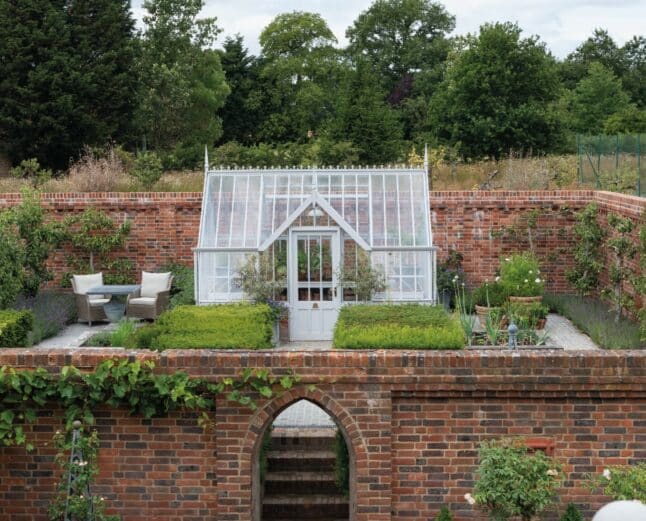 White victorian-style greenhouse within a walled garden. It has a flat-fronted lobby