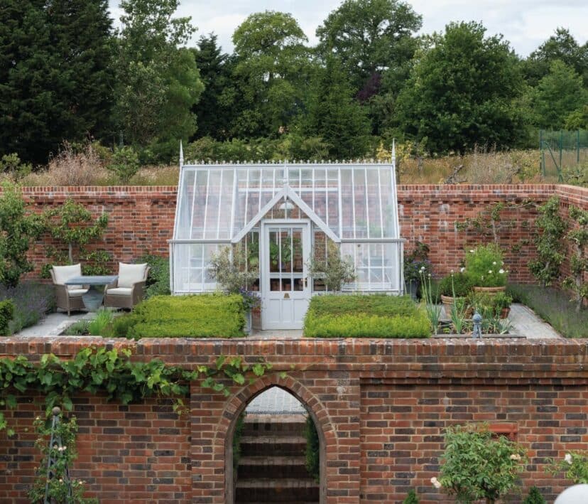 White victorian-style greenhouse within a walled garden. It has a flat-fronted lobby