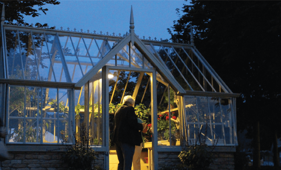 Victorian greenhouse at night with a single interior light. there are people in the greenhouse