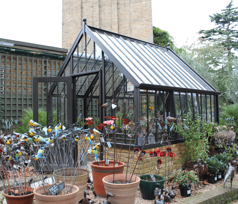 Black National Trust greenhouse on a sand brick base with potted plants in front.