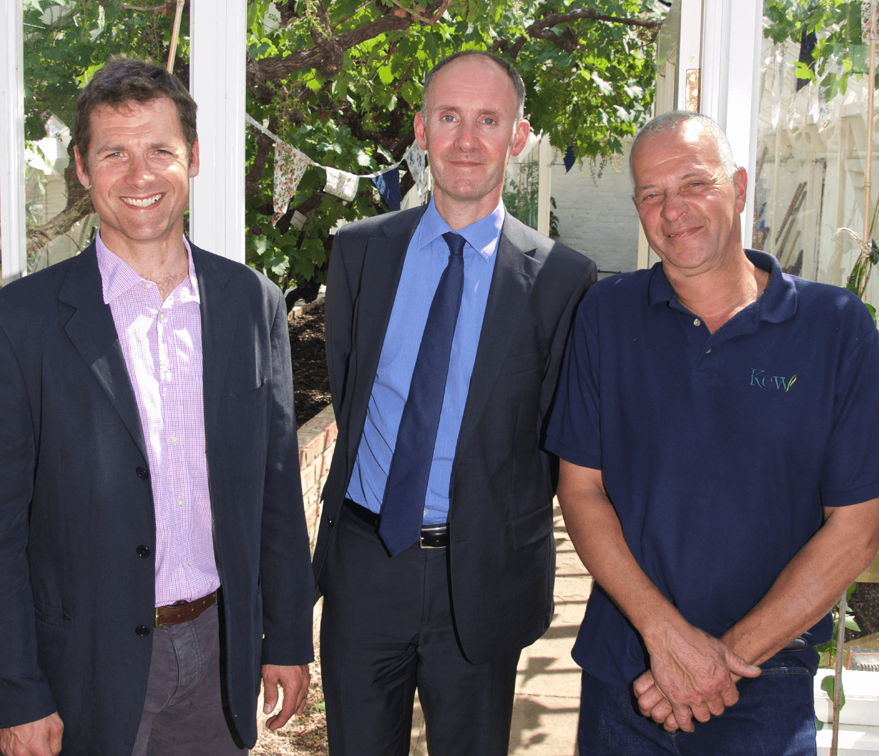 Three men smiling at the camera in front of a glasshouse