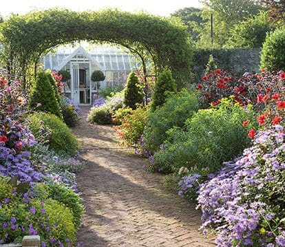 White Victorian greenhouse at the end of a stone path . The path is lined with colourful flowers and a foliage archway.