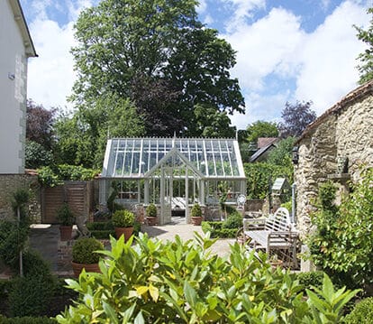 White aluminium greenhouse with a flat fronted lobby in a walled garden. There are several potted plants and shrubbery in front of the greenhouse.