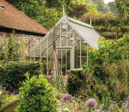 Small victorian greenhouse nestled behind greenery in a small garden.