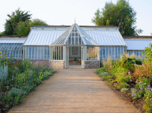 Stone path leading to large bespoke greenhouse in white with a hexagonal lobby on a brick base. The path is lined with plants and foliage.