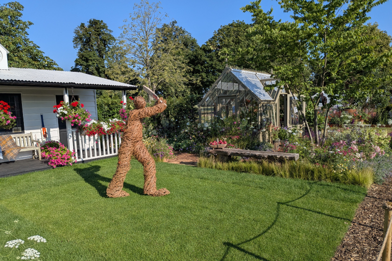 A handcrafted woodsage mottisfont Alitex greenhouse sits among abundant cottage-style planting, flowering perennials and mature trees in The Cricket Pavilion Garden at the RHS Badminton Flower Show.