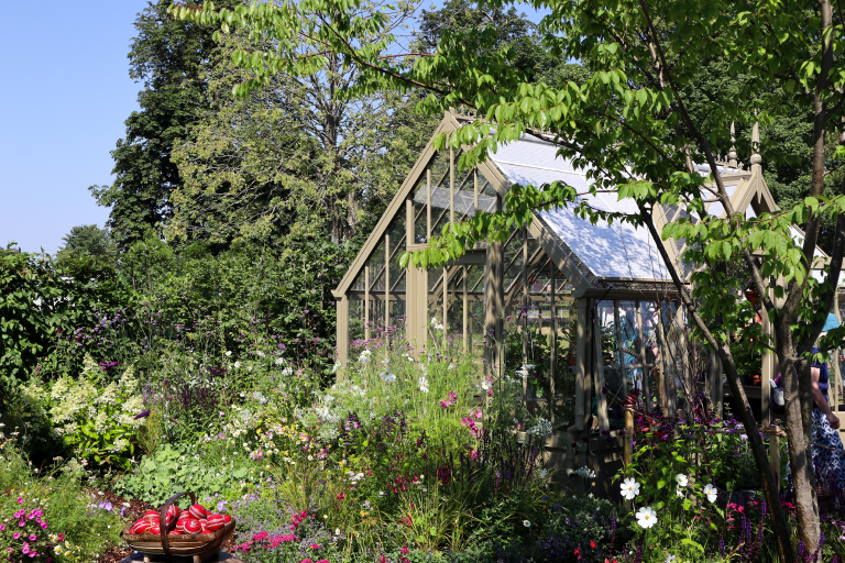 A handcrafted woodsage mottisfont Alitex greenhouse sits among abundant cottage-style planting, flowering perennials and mature trees in The Cricket Pavilion Garden at the RHS Badminton Flower Show.